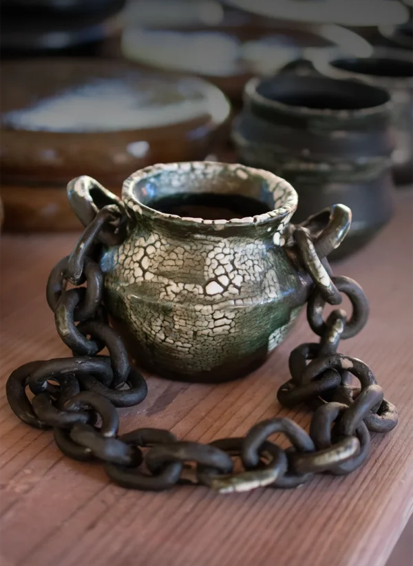 Pequeño jarrón de cerámica de alta temperatura con esmalte craquelado y cadena decorativa sobre mesa de madera.