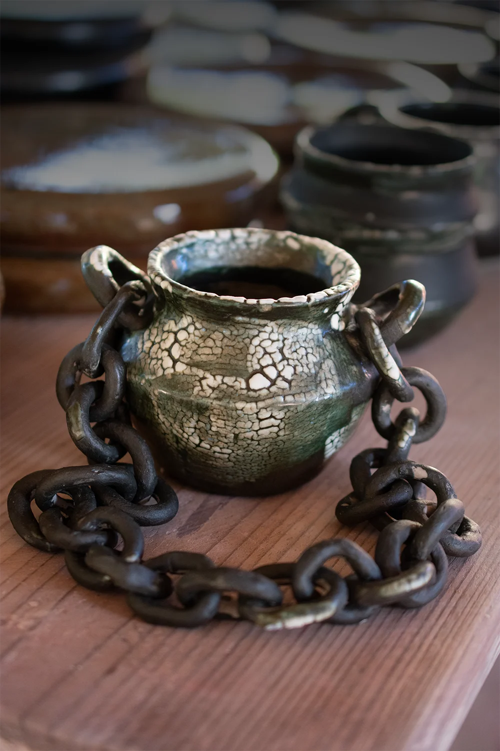 Pequeño jarrón de cerámica de alta temperatura con esmalte craquelado y cadena decorativa sobre mesa de madera.